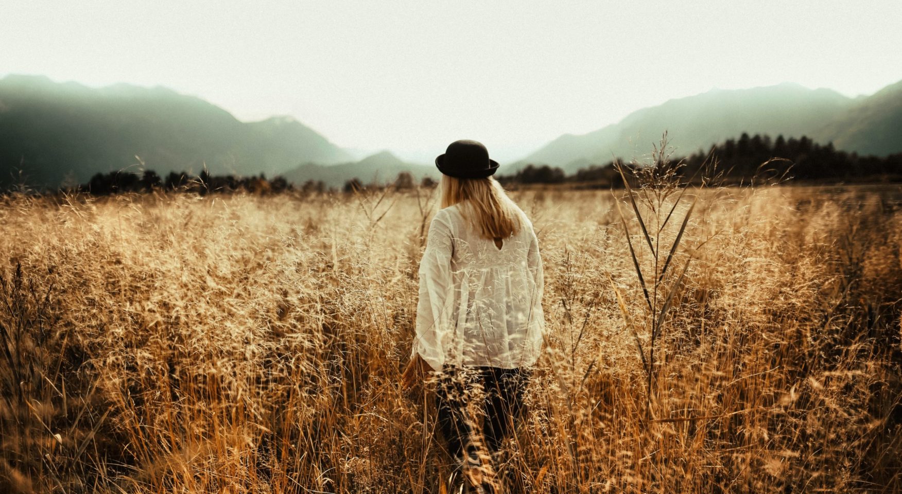 Woman wearing white lacy blouse and black hat walking through meadow of brown brush