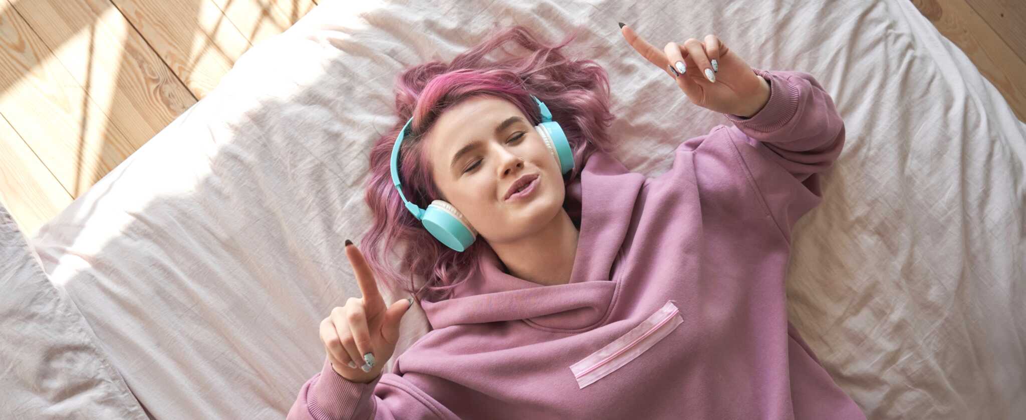 Happy funny teen girl with pink hair wear headphones lying in comfortable bed listening new pop music enjoying singing song with eyes closed relaxing in cozy bedroom at home. Top view from above.